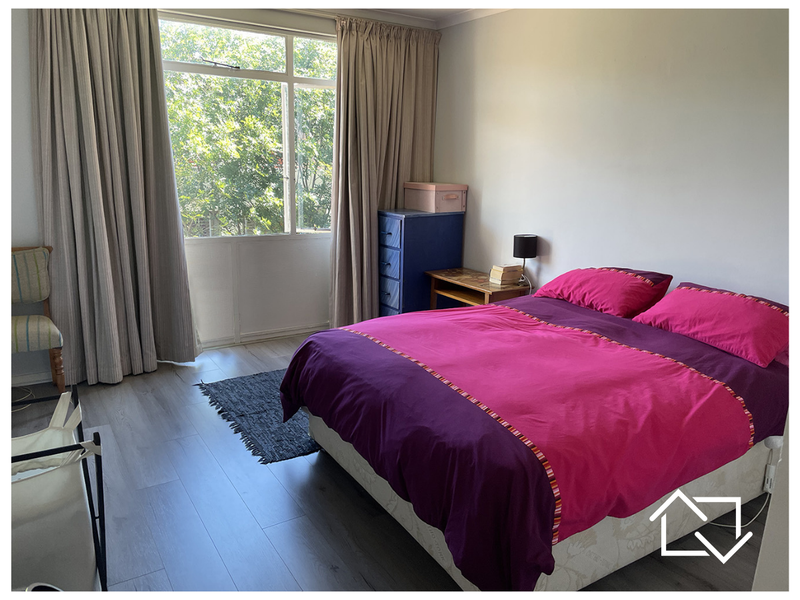 A well-lit bedroom featuring modern laminate flooring and a large window with floor-to-ceiling curtains, offering a bright and functional living space.