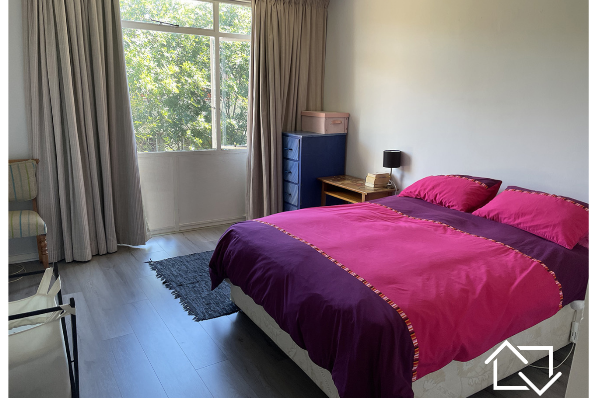 A well-lit bedroom featuring modern laminate flooring and a large window with floor-to-ceiling curtains, offering a bright and functional living space.