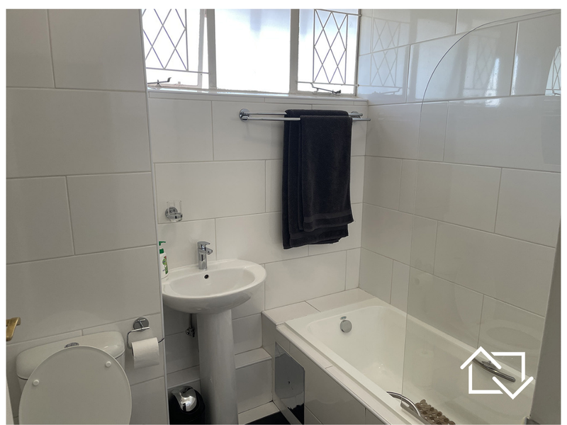 A clean, functional bathroom featuring white wall tiles, a pedestal basin, a toilet, and a bathtub with a glass shower screen.