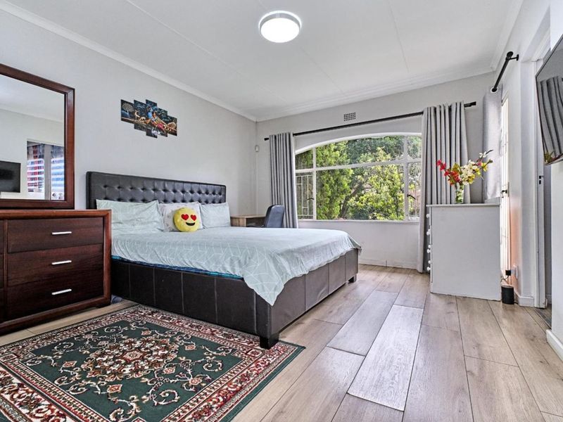 A well-lit bedroom featuring modern laminate flooring, neutral wall tones, and a large arched window providing natural light and garden views.