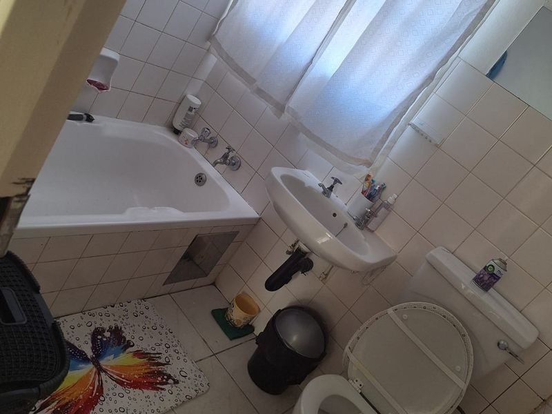 A compact bathroom featuring a bathtub, wall-mounted basin, and toilet with white tiled walls and flooring, requiring cosmetic updates.