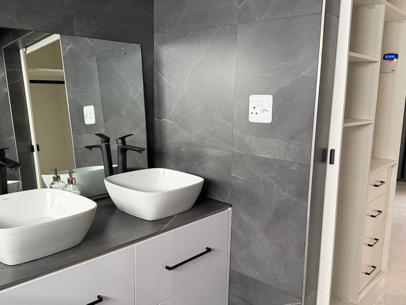 Modern bathroom featuring a double vanity with white vessel sinks, matte black fixtures, and dark grey marble-effect wall tiling, adjacent to built-in storage.