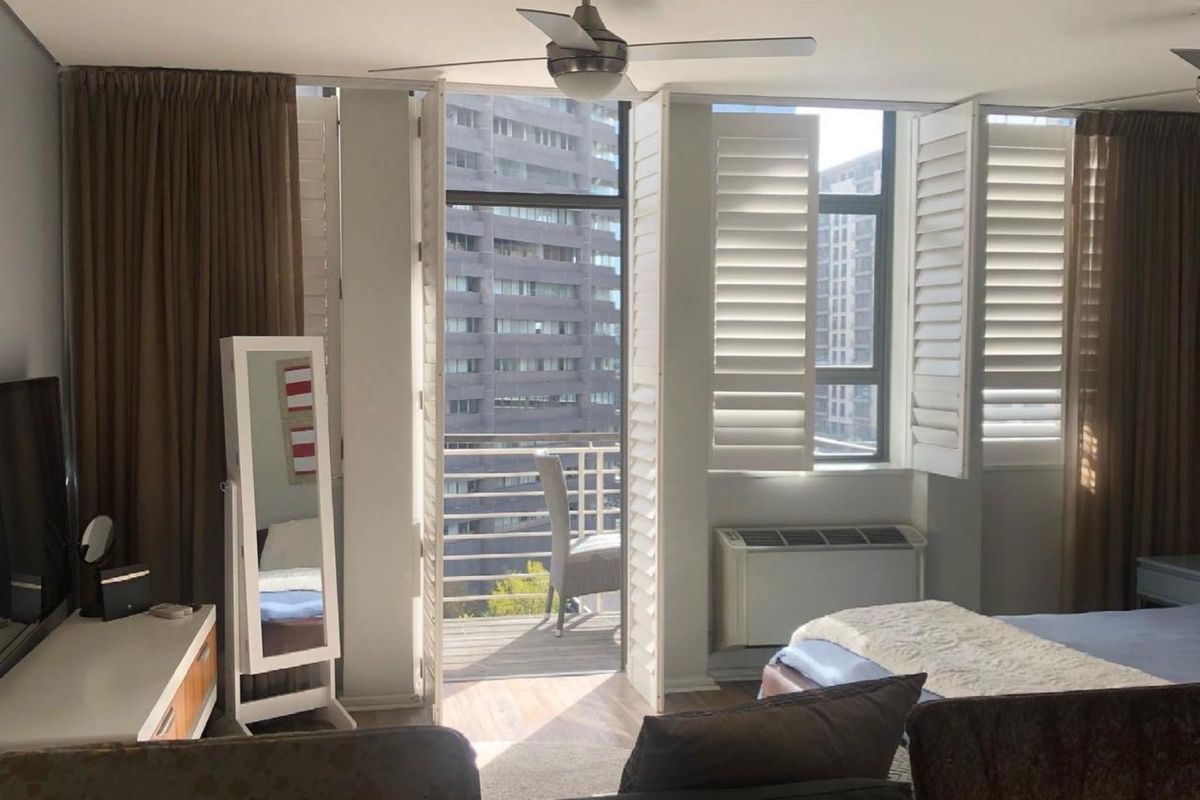 A bedroom featuring a ceiling fan, plantation shutters, and direct access to a private balcony with urban views.