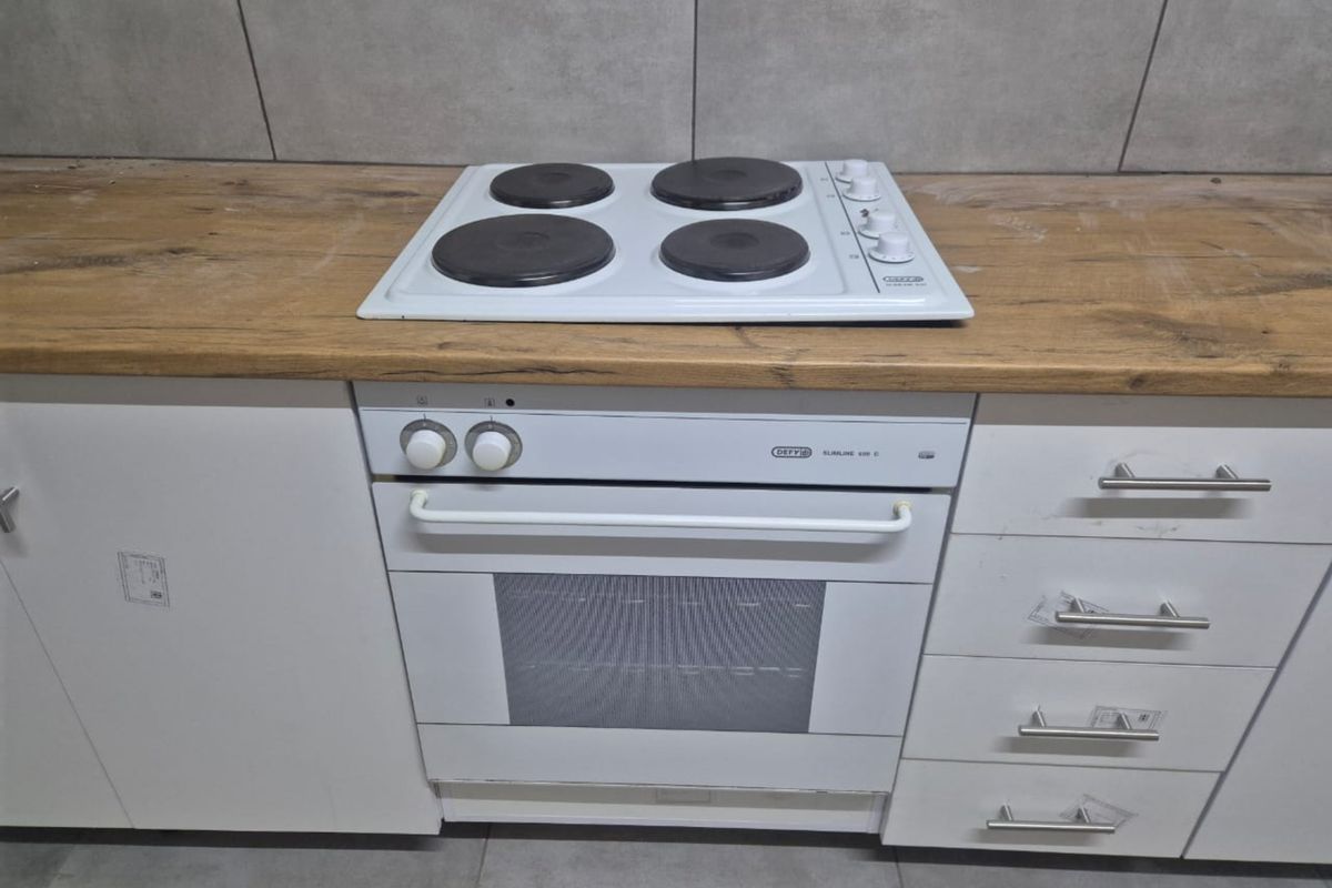 A kitchen section featuring a built-in white oven and electric hob set into a wood-look laminate countertop with tiled backsplash and white cabinetry.