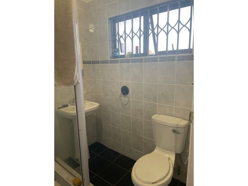 A functional bathroom featuring a pedestal basin, toilet, and shower enclosure with tiled finishes and a security gate on the window.