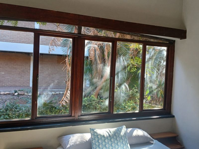 A bedroom featuring large timber-framed windows with garden views and a built-in headboard, offering a functional space with potential for cosmetic updates.
