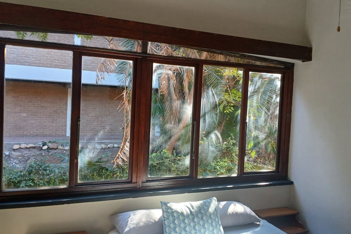 A bedroom featuring large timber-framed windows with garden views and a built-in headboard, offering a functional space with potential for cosmetic updates.