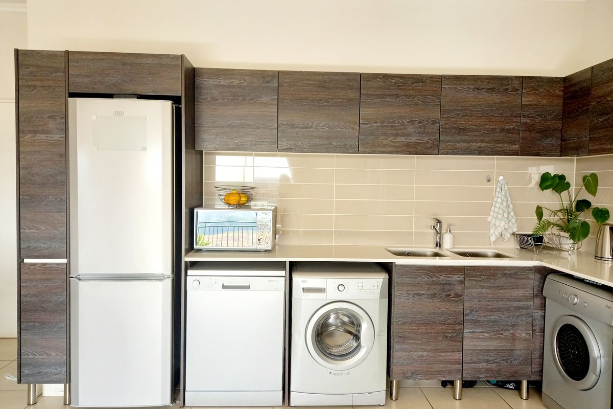 Modern kitchen featuring dark wood-grain cabinetry, tiled backsplash, double sink, and integrated space for multiple appliances including a dishwasher and washing machine.