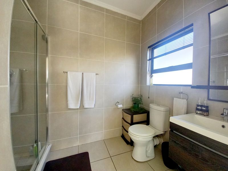 A well-maintained bathroom featuring a glass shower enclosure, modern vanity with integrated basin, neutral wall and floor tiling, and a window for natural light.