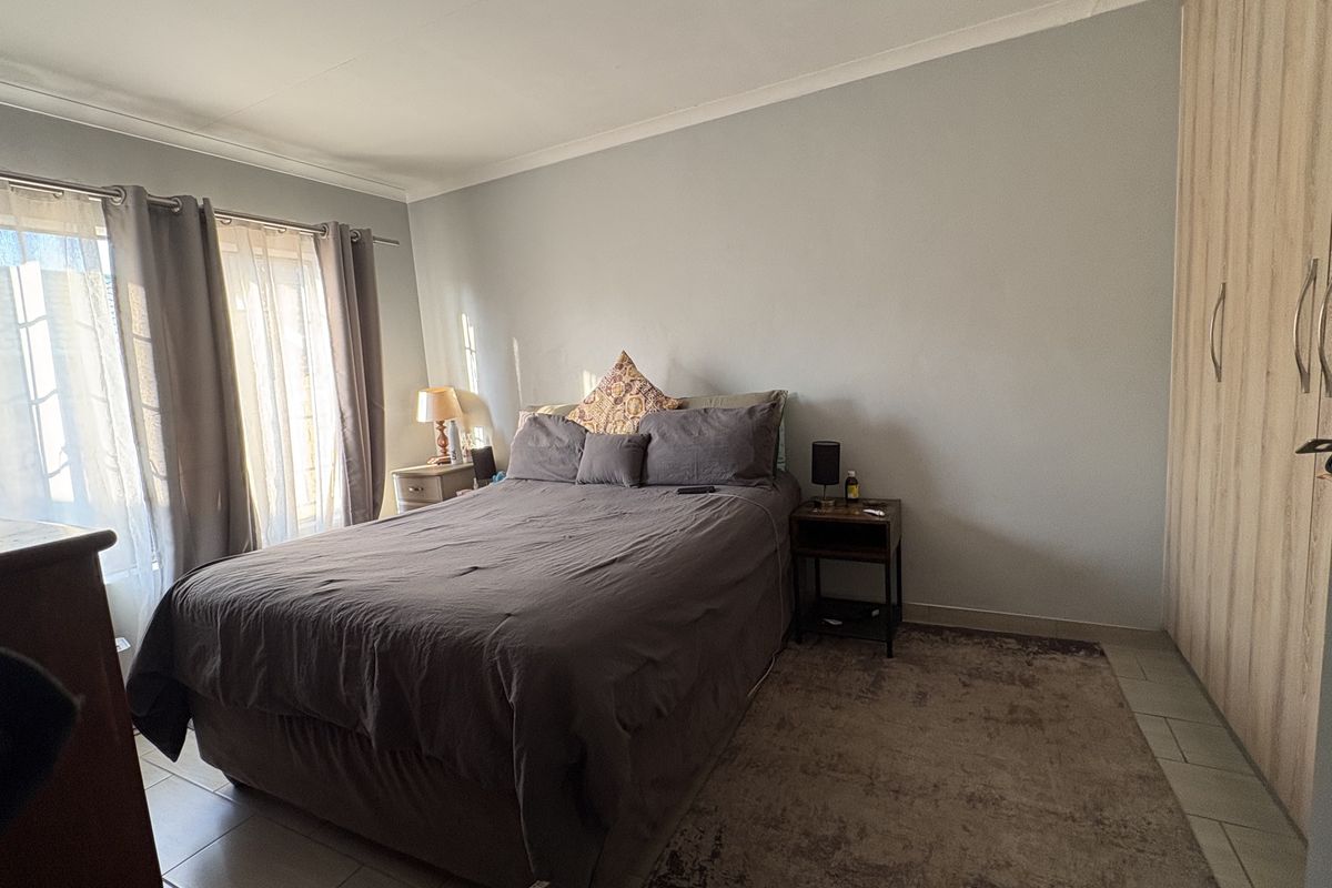 A well-lit bedroom featuring built-in wooden wardrobes, tiled flooring, and large windows with curtains, providing a functional and comfortable living space.