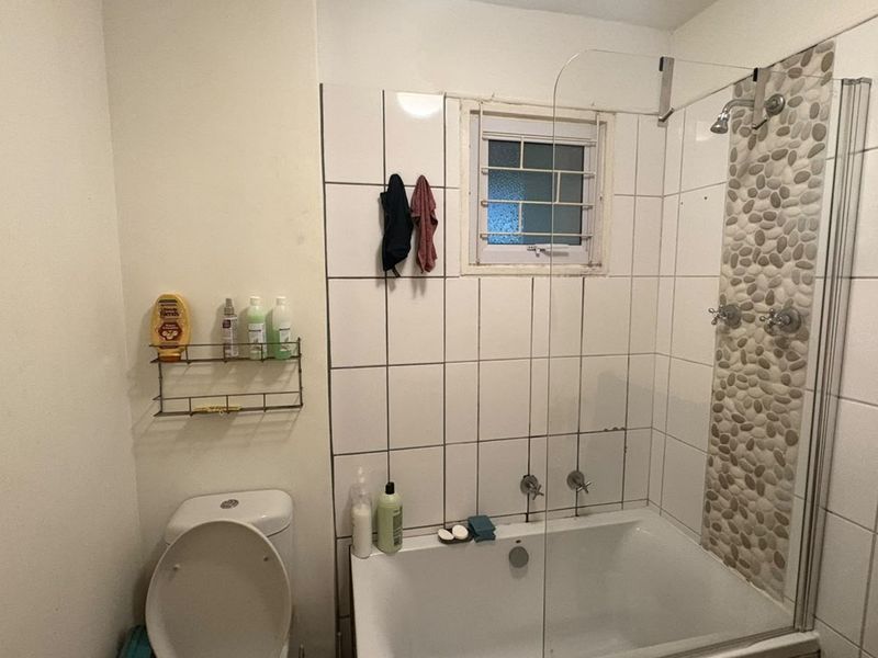 A functional bathroom featuring a combined bathtub and shower with a decorative pebble mosaic tile accent, white wall tiles, and a window with security bars.