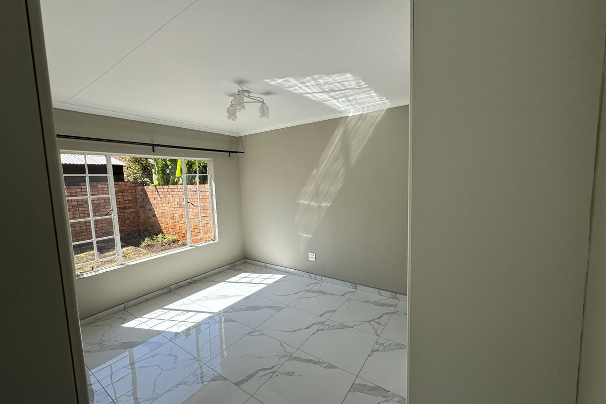 A bright, empty bedroom featuring modern marble-look floor tiles, neutral wall tones, and a large window overlooking the rear garden area.