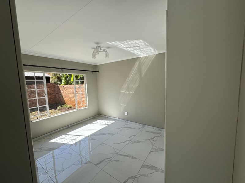 A bright, empty bedroom featuring modern marble-look floor tiles, neutral wall tones, and a large window overlooking the rear garden area.