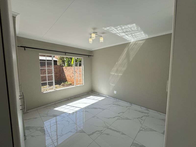 A bright, empty bedroom featuring polished marble-look floor tiles, neutral walls, and a window overlooking a private rear garden area.