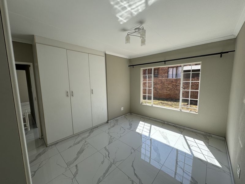 A bright, empty bedroom featuring modern marble-look tiled flooring, built-in cupboards, and a window with security bars overlooking the exterior.