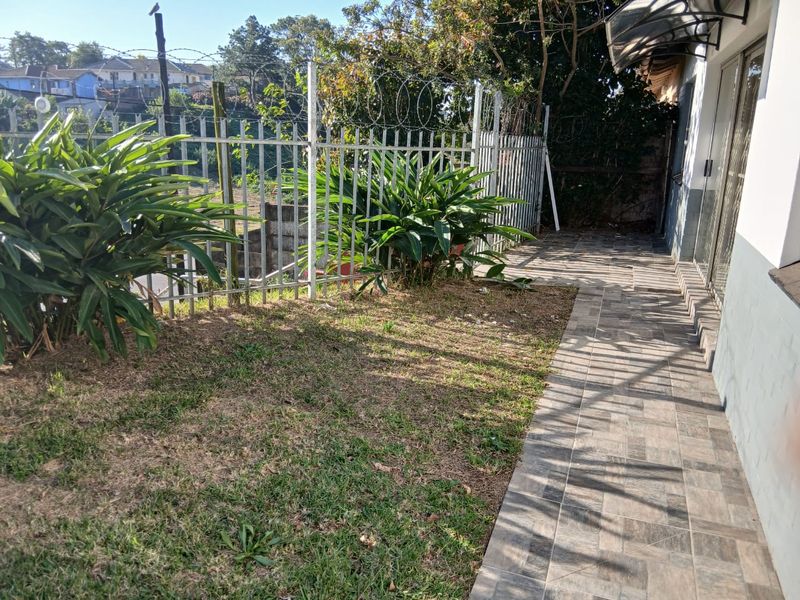 A rear exterior view of the cottage featuring a tiled patio area, metal security fencing with razor wire, and a small lawn.