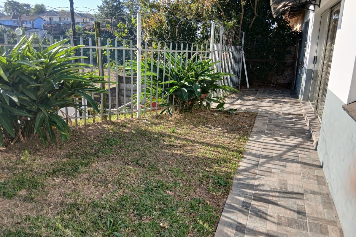 A rear exterior view of the cottage featuring a tiled patio area, metal security fencing with razor wire, and a small lawn.