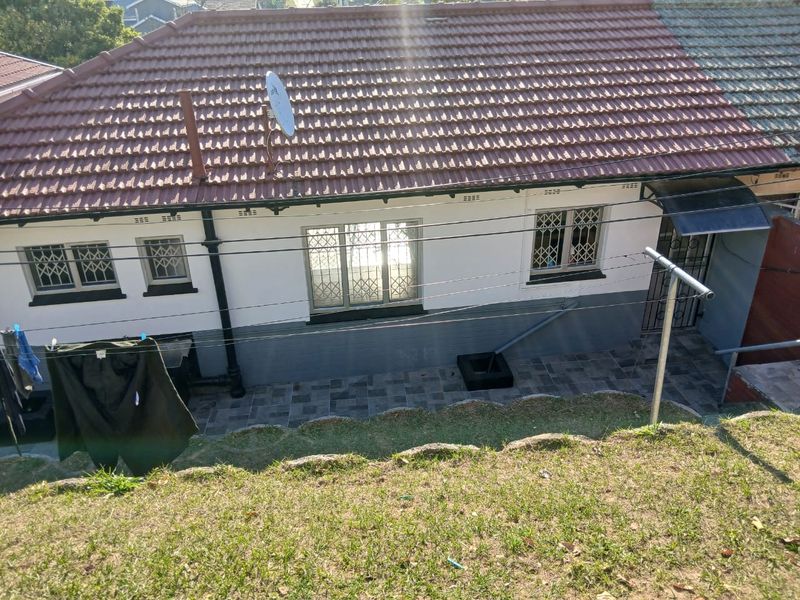 Rear view of the cottage featuring a tiled roof, security bars on windows, and a paved patio area adjacent to a sloped garden.