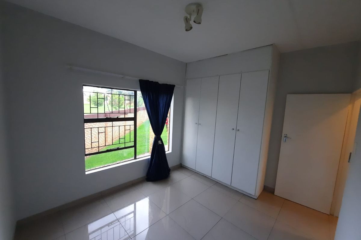 A bright, empty bedroom featuring neutral tiled flooring, built-in white cupboards, and a window with security bars overlooking the exterior.