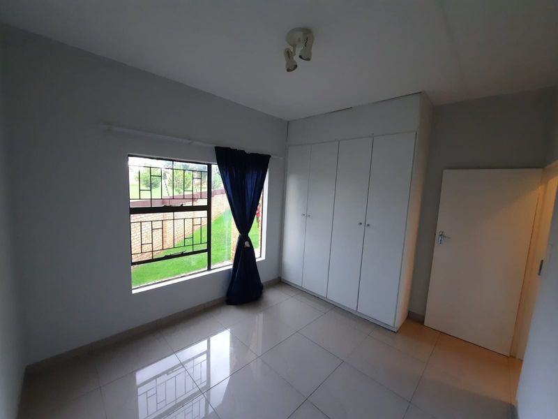 A bright, empty bedroom featuring neutral tiled flooring, built-in white cupboards, and a window with security bars overlooking the exterior.
