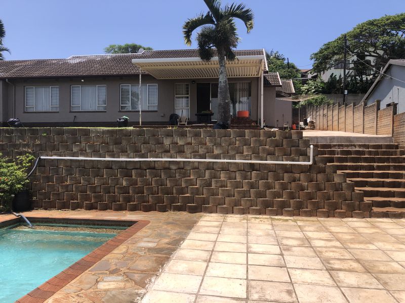 Rear exterior view featuring a swimming pool, paved patio area, and a tiered retaining wall leading up to the main house structure.