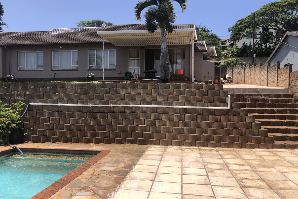 Rear exterior view featuring a swimming pool, paved patio area, and a tiered retaining wall leading up to the main house structure.