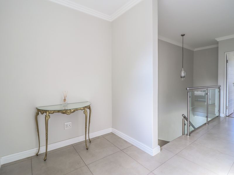 A bright and modern hallway featuring neutral tiled flooring, a sleek glass balustrade, and clean architectural lines leading to an adjacent room.