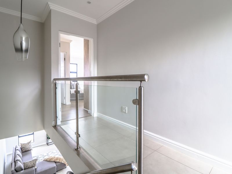 A bright, modern upstairs landing featuring a sleek glass balustrade with stainless steel handrails, tiled flooring, and neutral wall finishes.