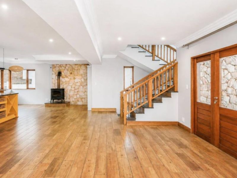 Spacious open-plan living area featuring a striking stone accent wall, wood-burning stove, and a classic wooden staircase with matching timber flooring.