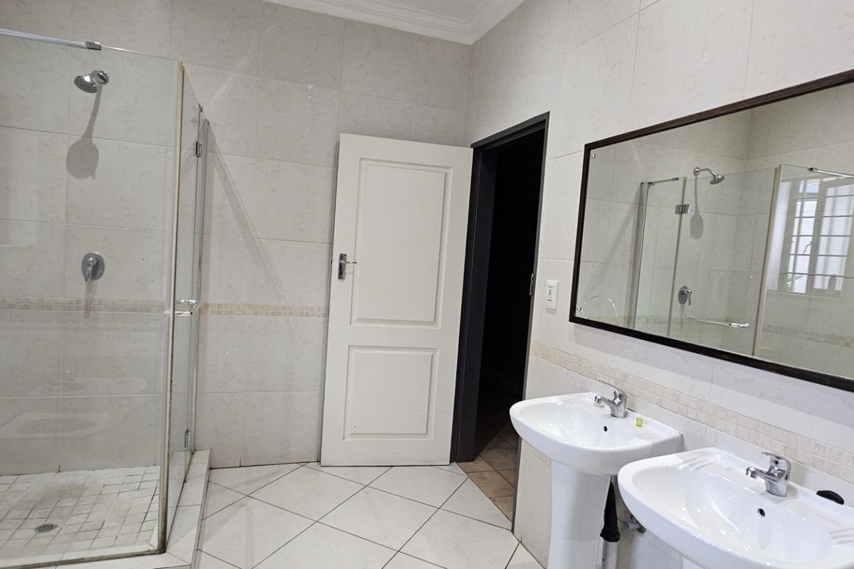 A clean, functional bathroom featuring a glass-enclosed shower, dual pedestal sinks, and neutral wall and floor tiling.