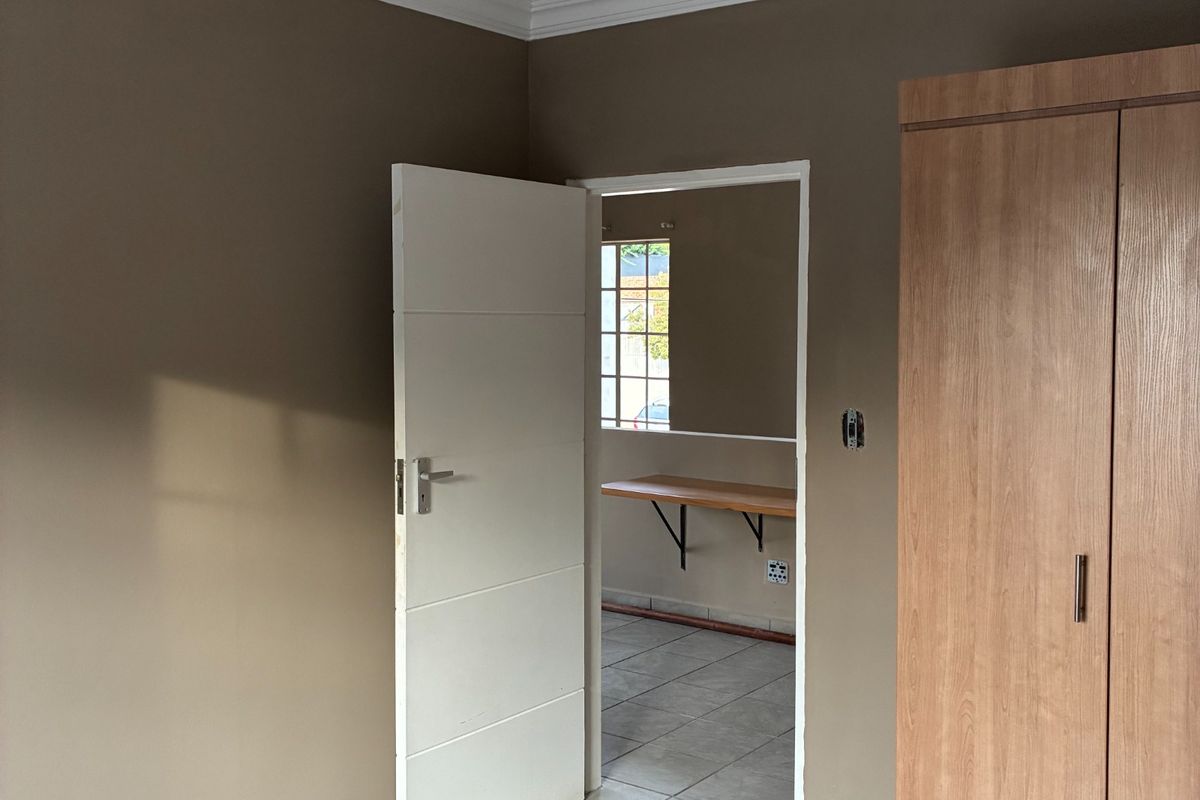 A clean, neutral-toned bedroom featuring tiled flooring, a built-in wardrobe, and decorative ceiling cornicing, with an adjacent room visible through the doorway.