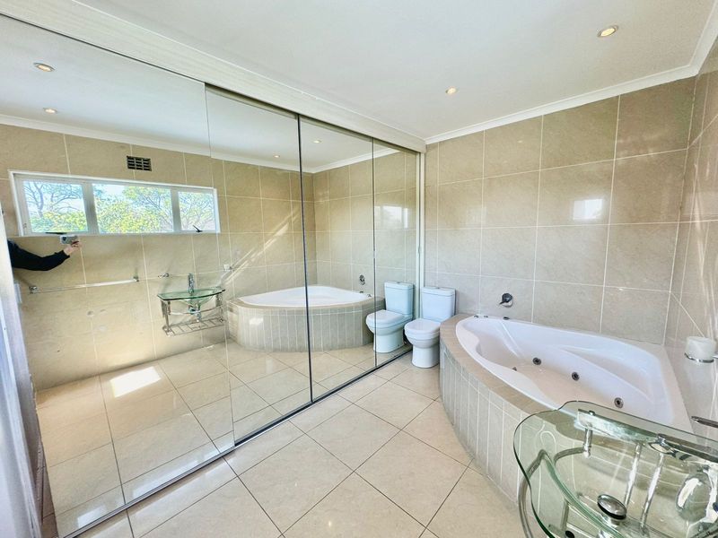 Spacious bathroom featuring a built-in corner spa bath, large wall-to-wall mirror, and neutral floor-to-ceiling tiling throughout.