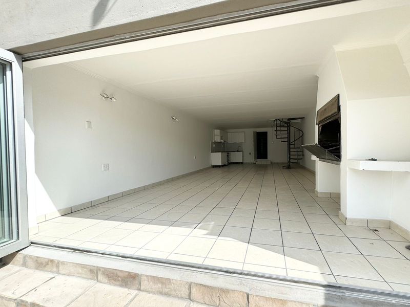 Spacious open-plan living area featuring tiled floors, a built-in braai, a spiral staircase, and a compact kitchen, opening out through large bi-fold doors.
