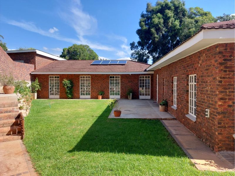 A well-maintained rear exterior view featuring a brick facade, tiled roof with solar panels, a concrete patio area, and a landscaped lawn.