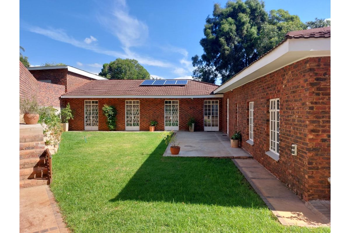 A well-maintained rear exterior view featuring a brick facade, tiled roof with solar panels, a concrete patio area, and a landscaped lawn.