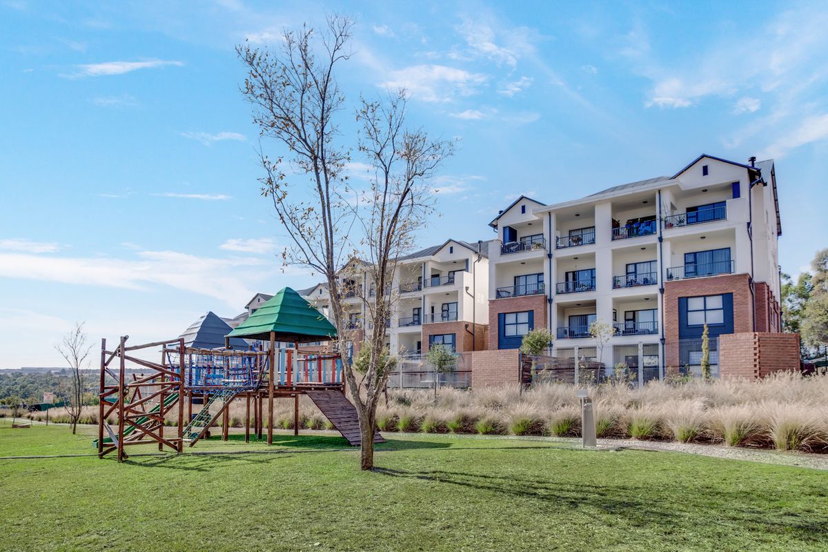 Exterior view of a modern multi-story apartment complex featuring private balconies, brick detailing, and an adjacent landscaped communal garden with a playground.
