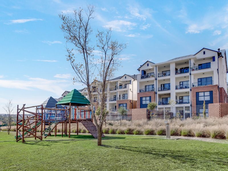 Exterior view of a modern multi-story apartment complex featuring private balconies, brick detailing, and an adjacent landscaped communal garden with a playground.