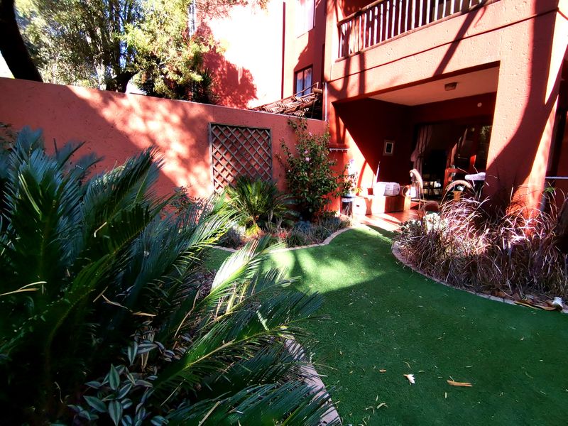 A private rear garden area featuring an artificial lawn, landscaped garden beds, a boundary wall with trellis, and a covered patio space.