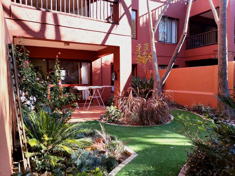 A private rear garden area featuring a covered patio, artificial lawn, and established landscaping, accessible via sliding glass doors from the apartment.