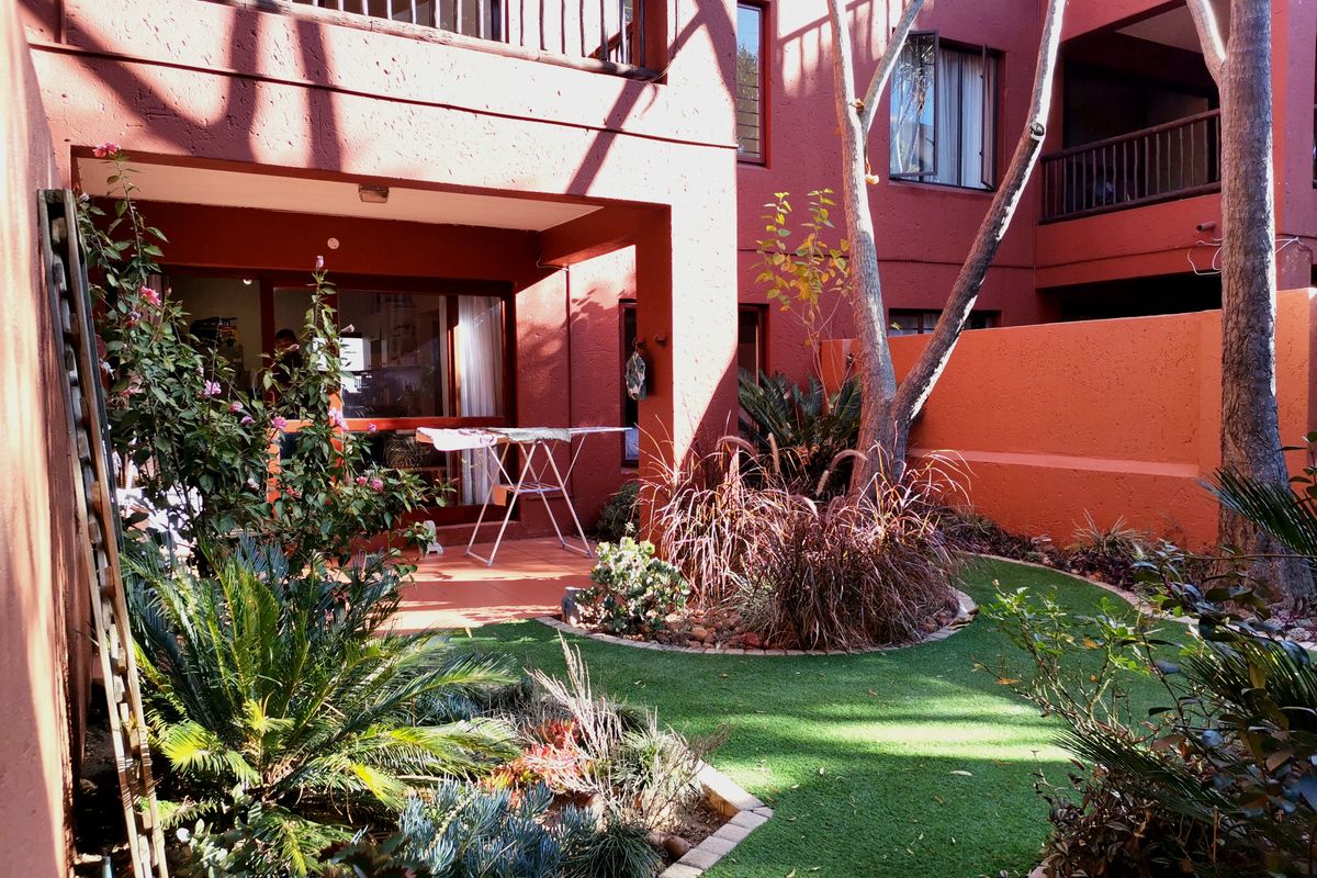 A private rear garden area featuring a covered patio, artificial lawn, and established landscaping, accessible via sliding glass doors from the apartment.