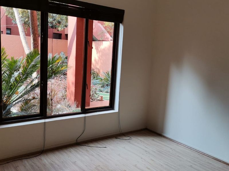 A bright, empty bedroom featuring light-toned laminate flooring and a large window with views of the exterior garden area.