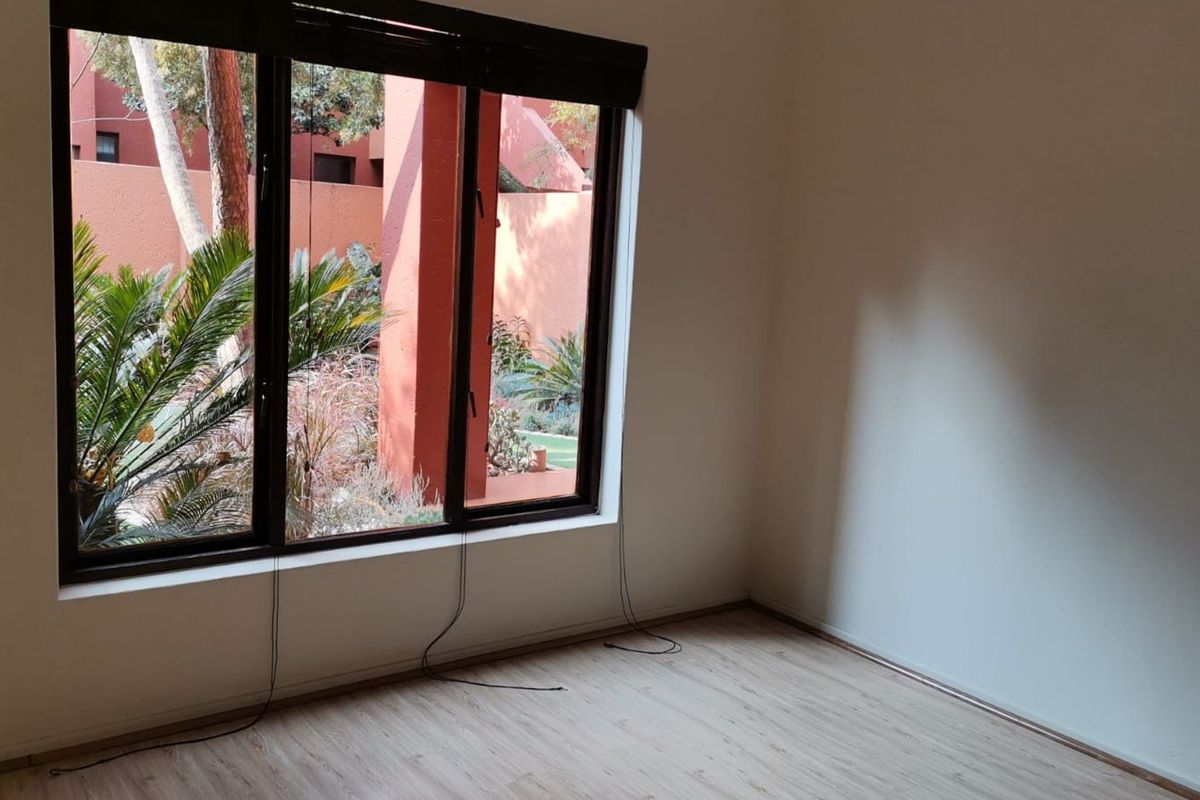 A bright, empty bedroom featuring light-toned laminate flooring and a large window with views of the exterior garden area.