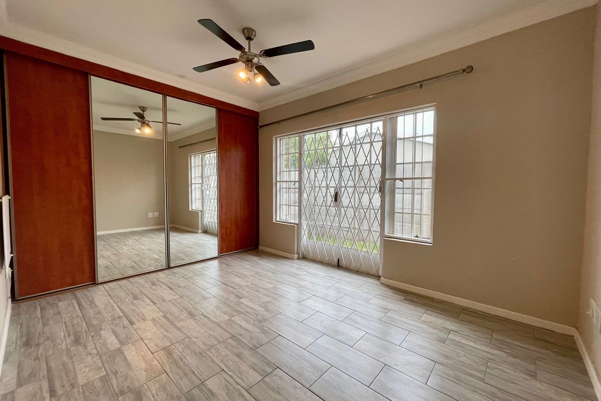 A bright, empty bedroom featuring modern wood-look tiled flooring, a large built-in mirrored wardrobe, a ceiling fan, and security gates on the window.