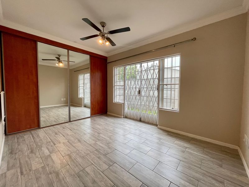 A bright, empty bedroom featuring modern wood-look tiled flooring, a large built-in mirrored wardrobe, a ceiling fan, and security gates on the window.