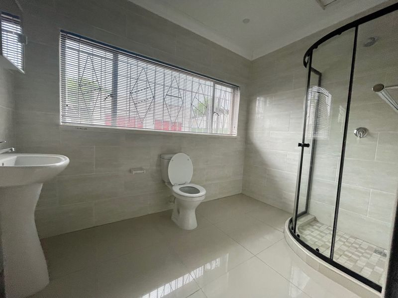 A clean, modern bathroom featuring a pedestal basin, a toilet, and a curved glass shower enclosure with neutral wall and floor tiling.
