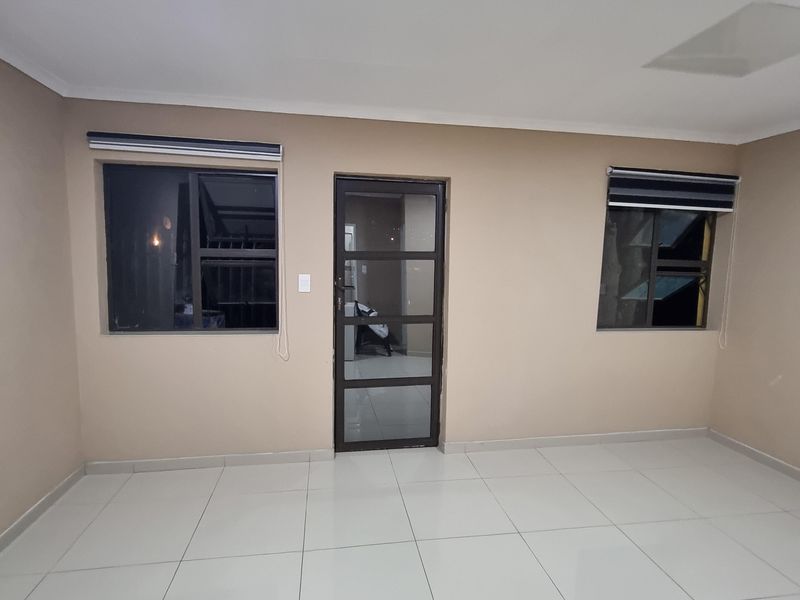A bright, empty living room featuring neutral tiled flooring, modern aluminium window frames, and fitted roller blinds.