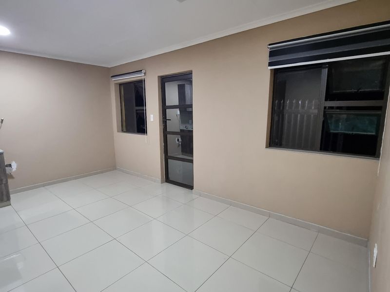A bright, empty living area featuring neutral wall tones, large format floor tiles, recessed lighting, and modern aluminium window frames with roller blinds.