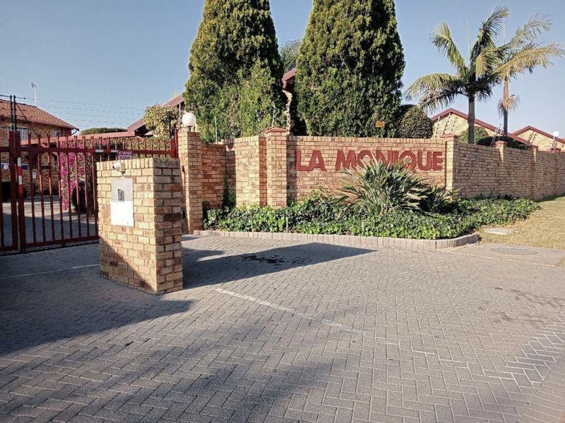 The entrance to La Monique townhouse complex featuring a brick boundary wall, paved driveway, and secure metal gate access.