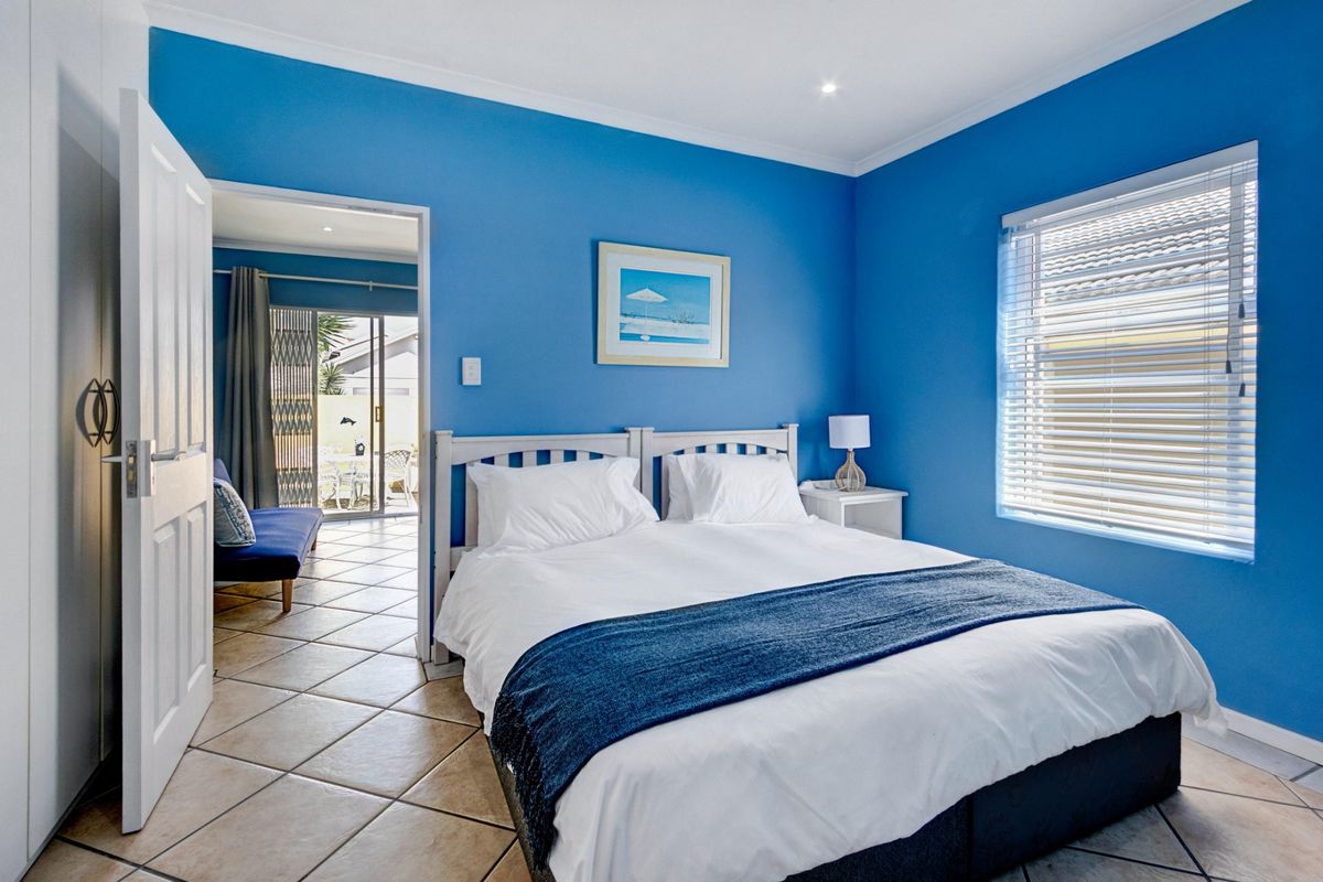 A bright bedroom featuring blue walls, tiled flooring, and a window with fitted blinds, with direct access to an adjoining living area.