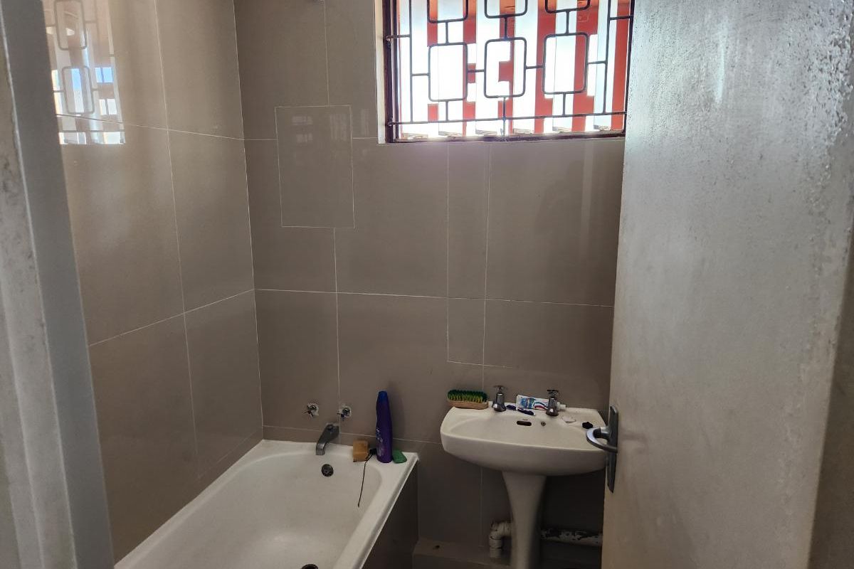 A functional bathroom featuring tiled walls, a bathtub with shower mixer, and a pedestal basin with security bars on the window.